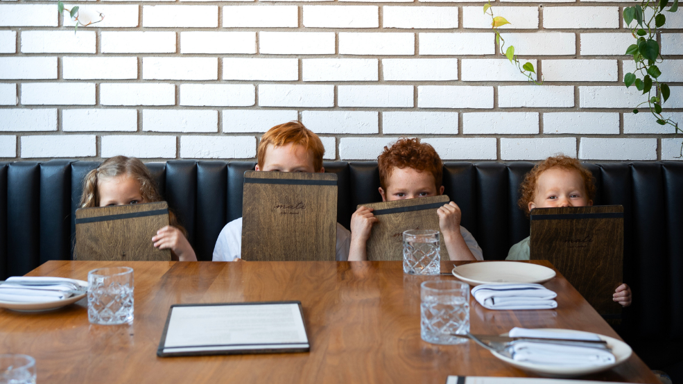 Four children sit in a restaurant booth, peeking out from behind wooden menus.