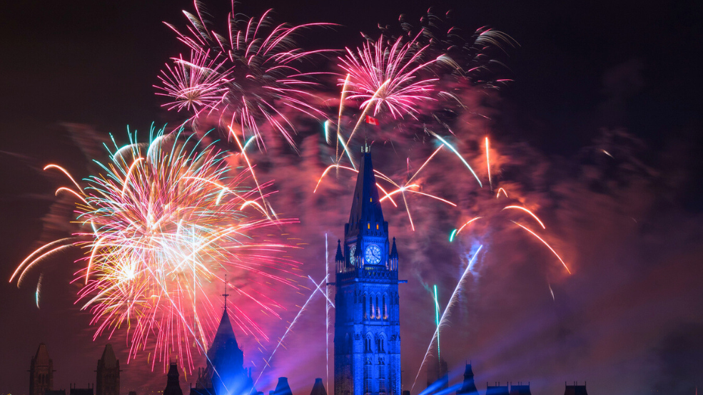 The Peace Tower is lit up in blue while red and pink fireworks explode behind it.