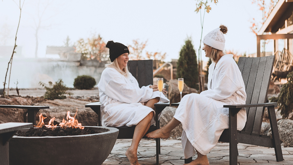 Two women in white bathrobes at an outdoor spa enjoying mimosas.