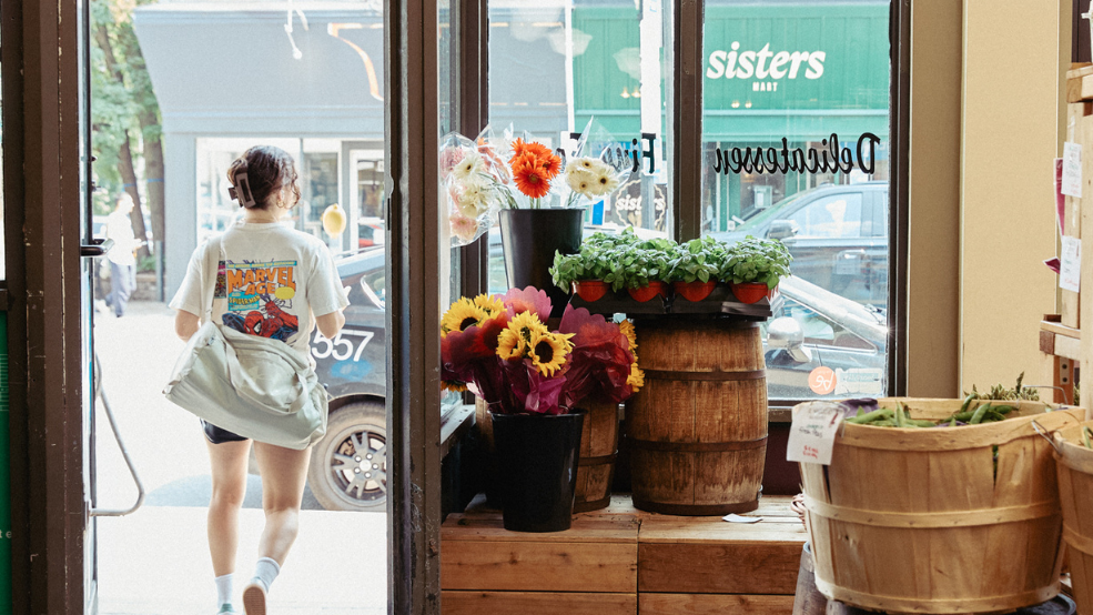 A person exits a shopfront full of plants and flowers looking onto a charming street.