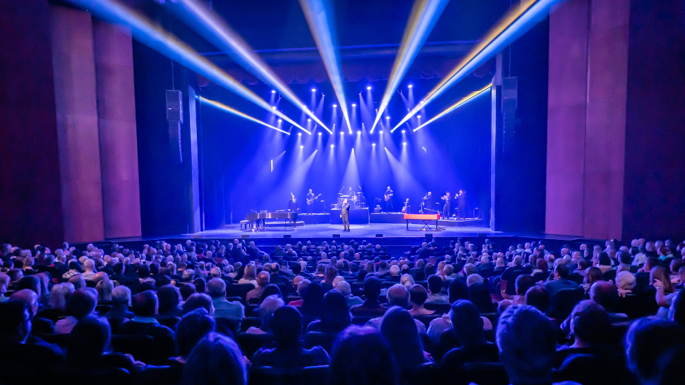 A 5 piece band performs on an indoor stage to a large audience.
