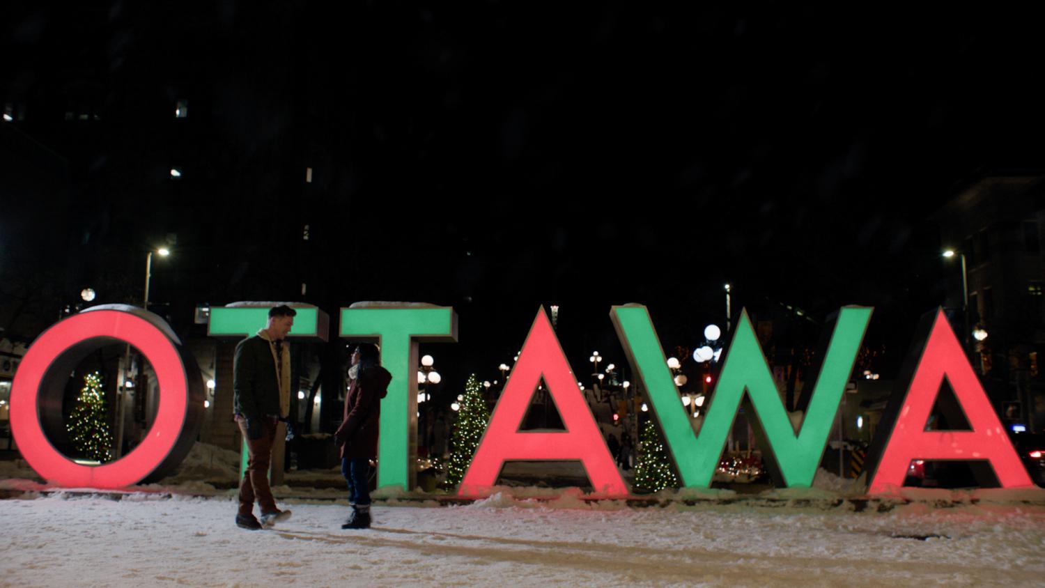 A couple standing in front of the Ottawa letters, lit in red and green. 