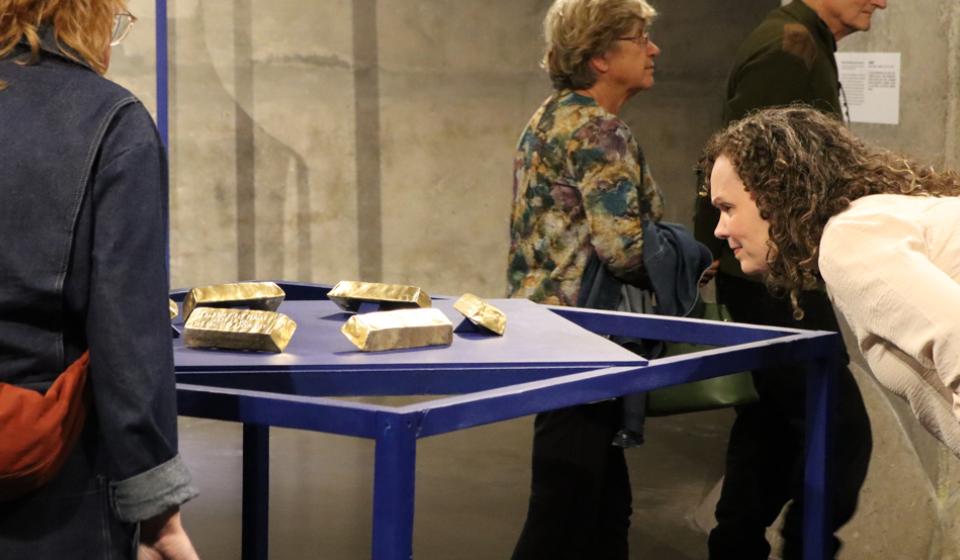 A person leans down to be eye level with a mixed media installation of gold bricks resting on a blue platform.