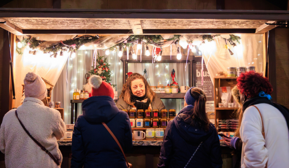 A market stand decorated with Christmas lights with several shoppers perusing.