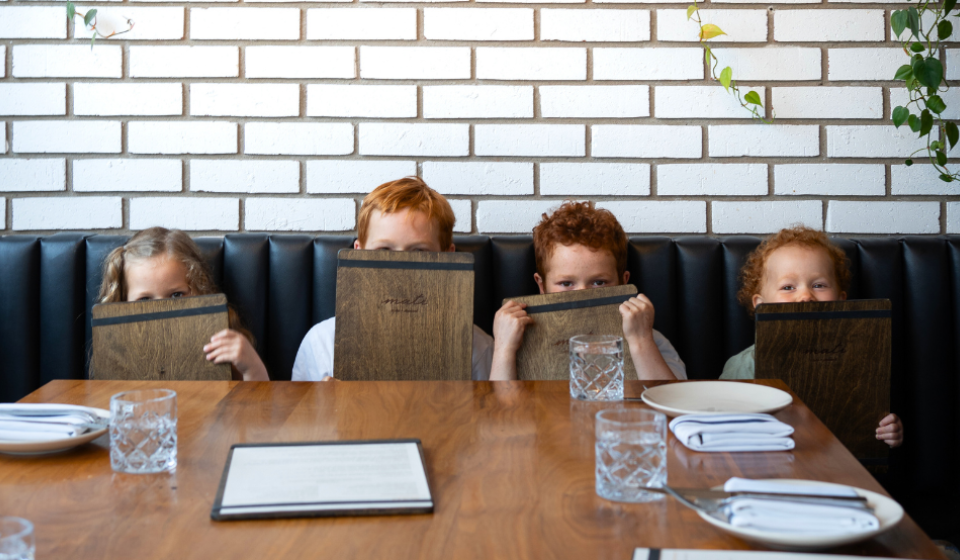 Four children sit in a restaurant booth, peeking out from behind wooden menus.