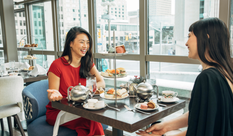Two people dressed formally sitting across a table with a three-tier tea tray filled with sweet and savoury fare.