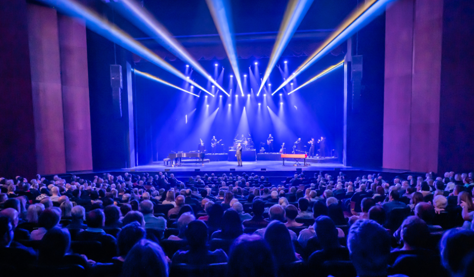A 5 piece band performs on an indoor stage to a large audience.