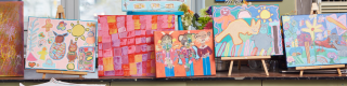 A photo of row of colorfully vibrant acrylic paintings on easels sitting on a shelf.