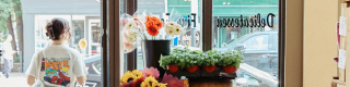 A person exits a shopfront full of plants and flowers looking onto a charming street. 