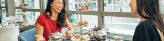 Two people dressed formally sitting across a table with a three-tier tea tray filled with sweet and savoury fare. 