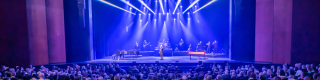 A 5 piece band performs on an indoor stage to a large audience. 
